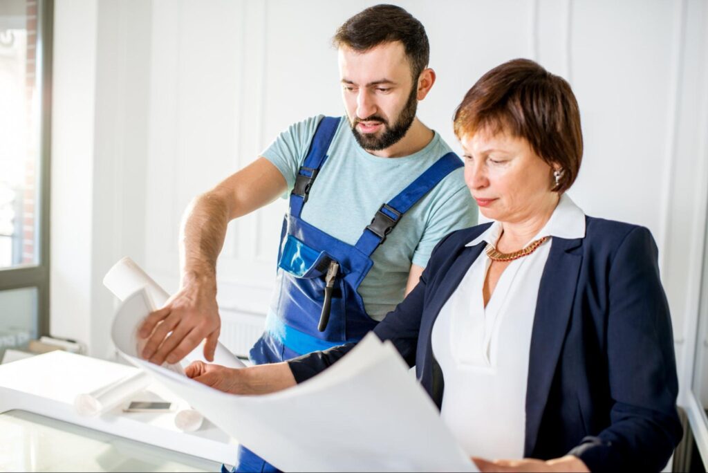 A bearded bathroom remodeling contractor in denim overalls discusses the project, referring to the blueprint with a female client in a corporate suit.