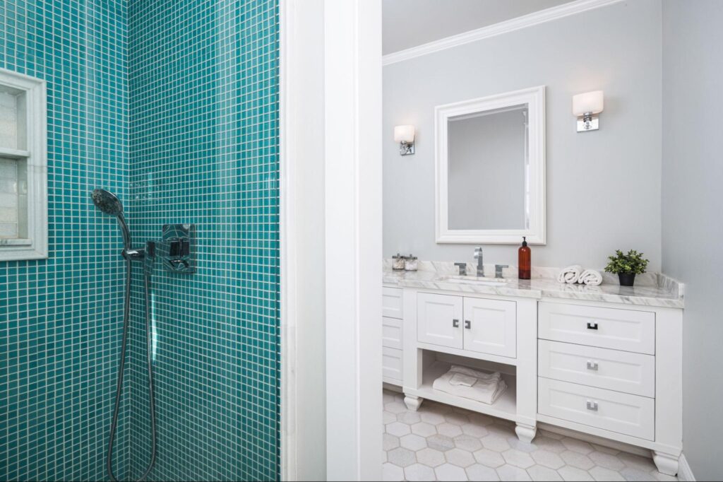 A spacious bathroom in a newly constructed home features blue tiles complementing a white theme. A stainless steel shower, white walls, cabinets, a mirror, towels, and toiletries are visible in the background.